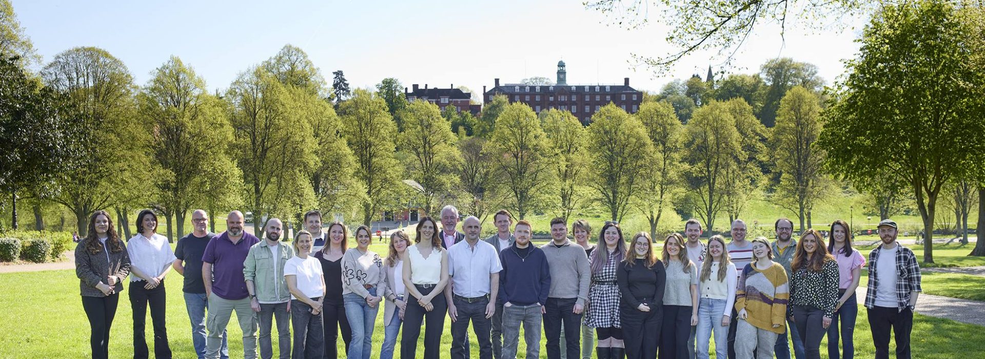 A team photo of the Clear and Buy-From Agencies standing together. It is a sunny day and they are in a park with trees behind them